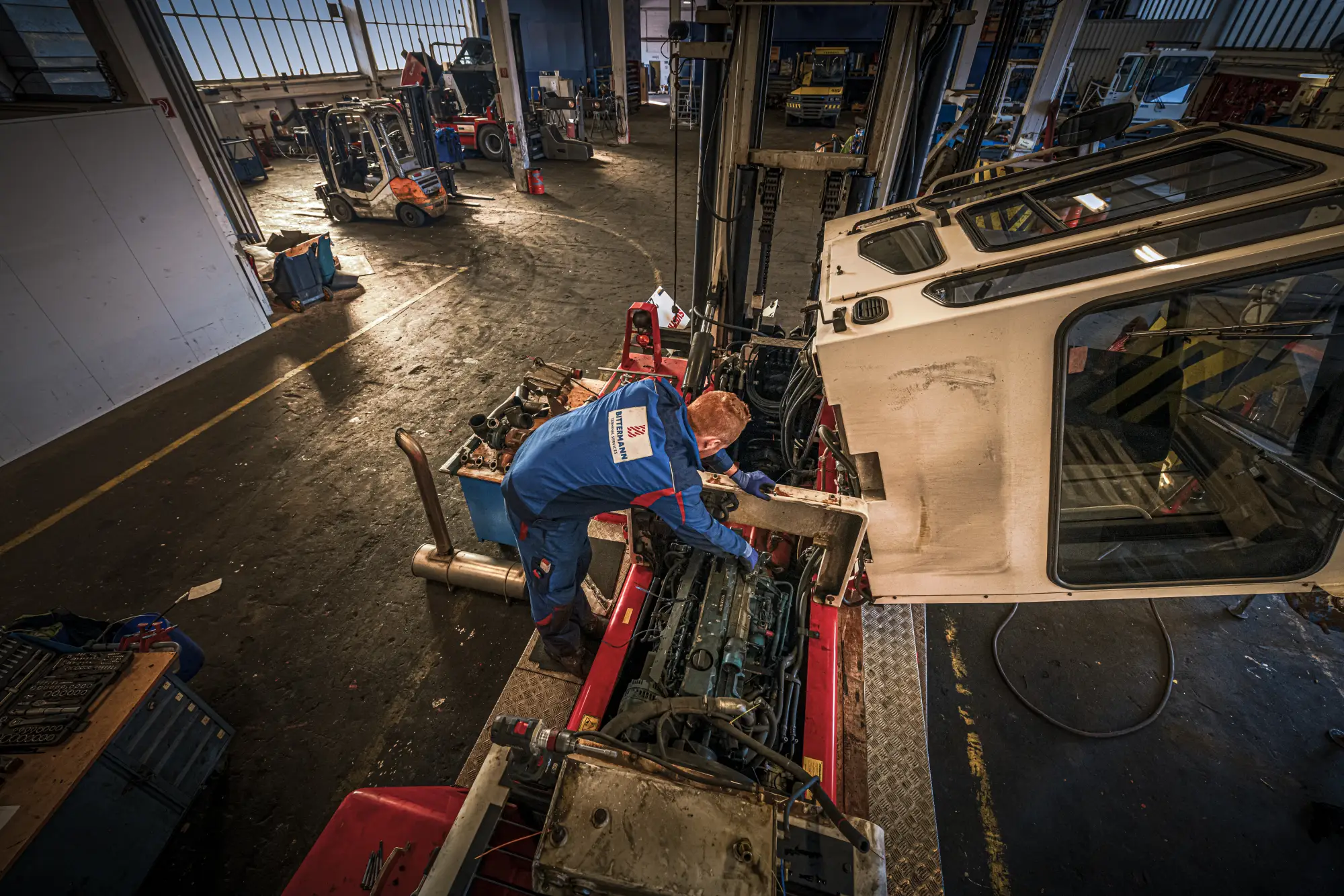 Mechaniker in blauem Overall arbeitet in der Werkstatthalle am Motor einer Maschine mit abgenommener Fahrerkabine